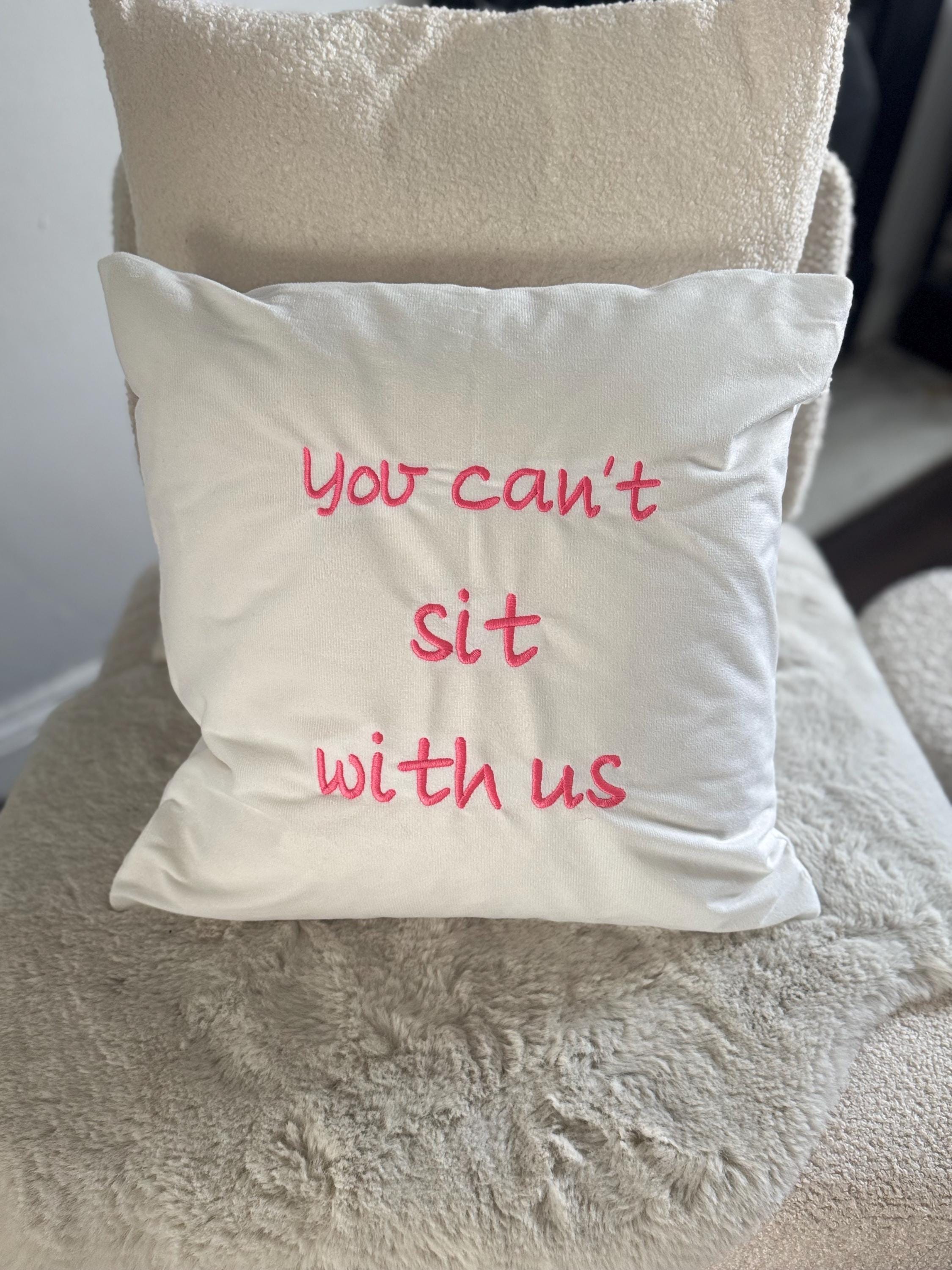 White cushion cover, you can’t sit with us logo embroidered in pink.