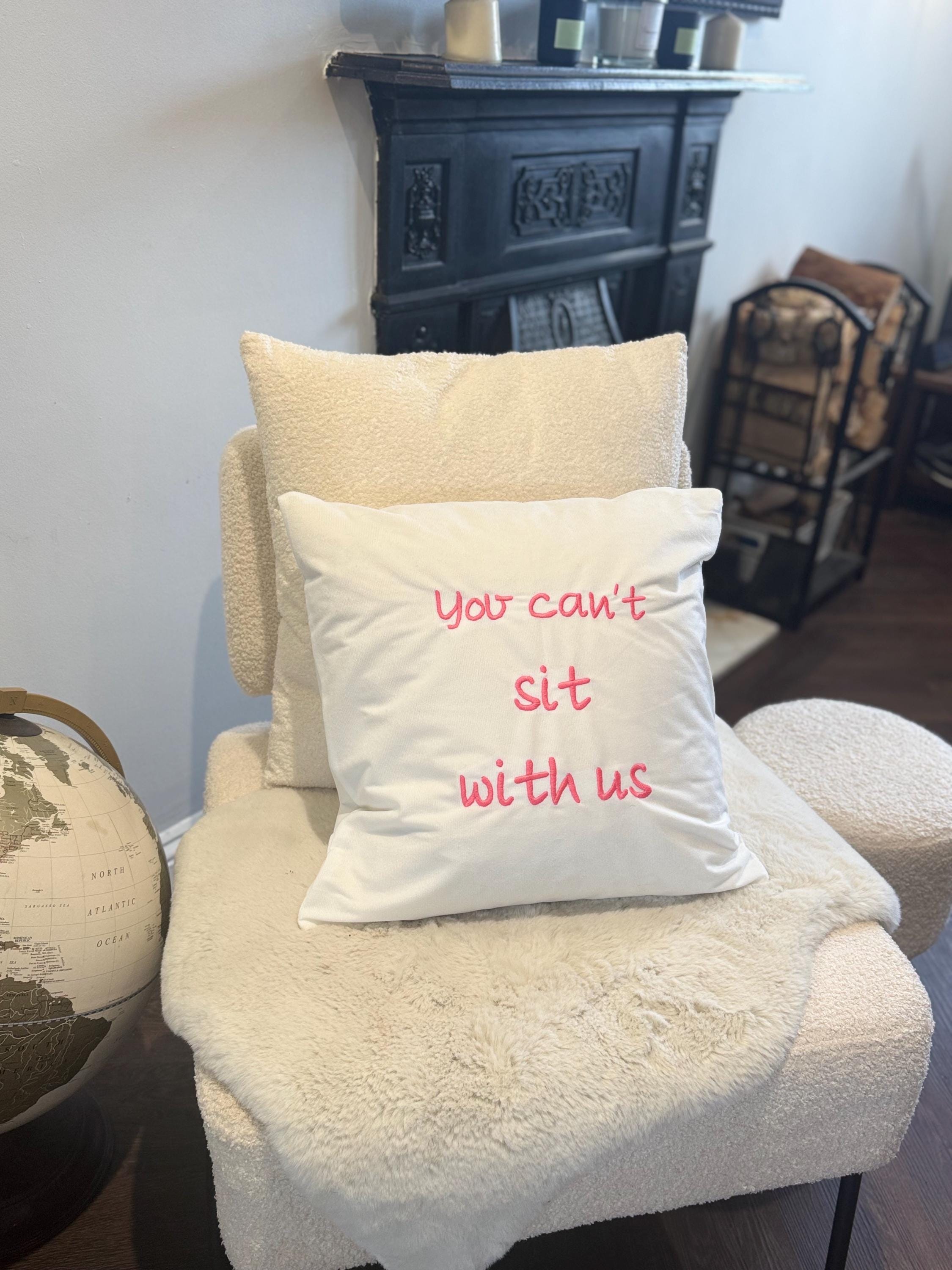 White cushion cover, you can’t sit with us logo embroidered in pink.